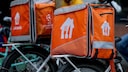 Several orange Lieferando delivery bags with white logos and gray reflector stripes are mounted on the luggage racks of bicycles. One of the bags also features the UEFA Champions League logo. In the blurred background, there are several people, including one person in a yellow jacket.