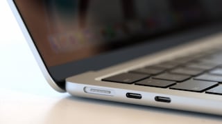 Close-up of the left side of a silver-colored laptop. Visible are the keyboard, the edge of the open screen, and two USB-C ports on the side of the casing. Next to them is a magnetic charging port with five contact points. The background is blurred, and the device rests on a light-colored surface.