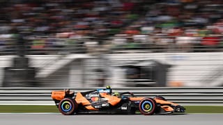 An orange and black Formula 1 race car from McLaren is seen from the side on a racetrack. The vehicle speeds along gray asphalt, adorned with several sponsor logos on its body and rear wing. The tires feature colored markings on the rims. In the background, grandstands filled with numerous spectators appear blurred due to the car's motion and a long exposure time. Guardrails and track boundaries run in front of the stands.
