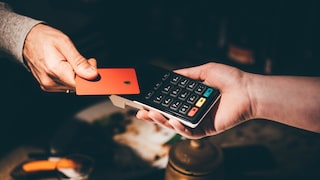 Close-up of a contactless card payment. One hand holds an orange card over a black card reader, which is held by another hand. The terminal features a keypad along with green, yellow, and red function buttons. The background is dark and blurred.