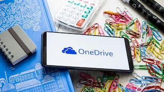 A smartphone displaying the OneDrive logo is placed on a desk. Surrounding the device are a blue notebook, a small spiral notepad, a calculator, several felt-tip pens, and numerous colorful paper clips in red, yellow, green, blue, and white. The screen shows the OneDrive logo and the word "OneDrive" against a white background.
