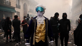 A person dressed in historical attire, wearing a white curly wig, a gold vest, and a dark blue coat, stands in the middle of a street. Their face is obscured by a gray cloth and large, blue mirrored protective goggles. In the background, there are numerous people, some wearing black face masks. The street is littered with trash and shrouded in thick smoke or fog.