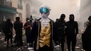 A person dressed in historical attire, wearing a white curly wig, a gold vest, and a dark blue coat, stands in the middle of a street. Their face is obscured by a gray cloth and large, blue mirrored protective goggles. In the background, there are numerous people, some wearing black face masks. The street is littered with trash and shrouded in thick smoke or fog.