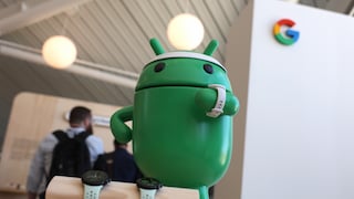 A large green Android figure stands at a trade show booth, wearing a white smartwatch on its arm. In front of the figure, two more smartwatches with light-colored bands rest on a wooden stand. In the background to the right, the multicolored Google logo is visible, while several blurred individuals are on the left.