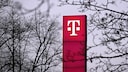 Tall, pink-colored stele with a white Telekom logo against a gray sky. Surrounding the stele are numerous bare tree branches, some of which are blurred in the foreground.