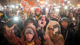 A large crowd gathers at night in a brightly lit square in front of historic buildings. Many people hold up smartphones with their flashlights on. In the center of the image, an orange heart with the white inscription "OSTRAVA" is visible. Several people are wearing rain jackets, hats, and hoods. In the background, a Czech flag is being held up.