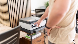 A person stands in front of a printer in an office, touching the device's illuminated display with a finger. The printer is placed on a small cabinet with drawers. In the background, wooden wall panels, desks, and office chairs are visible. The image primarily focuses on the printer, the person's arm, and the control panel of the device.