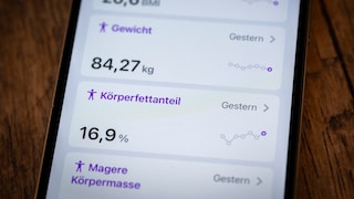 Close-up of a smartphone on a wooden surface. The display shows health data, including a BMI of 26.6, a weight of 84.27 kilograms, and a body fat percentage of 16.9 percent. Small trend graphs are visible next to the values.