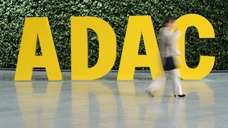 The image features large yellow letters spelling "ADAC" against a dense green plant wall. In the foreground, a person walks through the scene, appearing noticeably blurred due to motion. The lettering and parts of the person are reflected on the smooth floor.