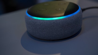 The image shows a compact, round smart speaker with a dark gray fabric cover and a black top. A blue-turquoise light ring glows at the upper edge. Several control buttons are visible on the top. The speaker is placed on a dark surface, with the background blurred and also dark.