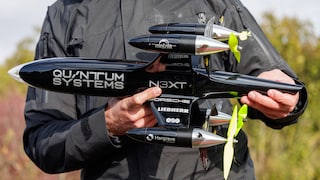 A person holds a black drone with multiple propellers in their hands. The body of the drone features the names Quantum Systems and N3XT, with vegetation visible in the background.