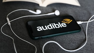 A smartphone with the Audible logo lies next to white headphones and an open book.