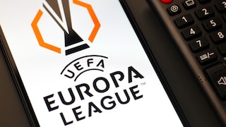 In the photo, a smartphone is placed next to a black TV remote control. The bright display of the smartphone prominently shows the black-and-orange UEFA Europa League logo. To the right, the red power button and the number keys of the remote are visible, among other features.