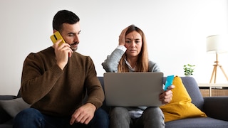 Two people are sitting on a sofa in front of a laptop. One person is on the phone with a focused expression, while the other is looking at the screen with a furrowed brow, holding a bank card in hand.
