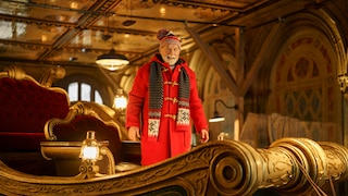 In the image, a person stands in an elaborately designed, Christmas-themed sleigh. The person is wearing a long red coat, a red patterned hat, and a black, red, and white scarf. The sleigh is adorned with gold-colored embellishments, dark surfaces, and red velvet. Several glowing lanterns provide warm light. In the background, decorated walls, arches, and a mirrored ceiling are visible.