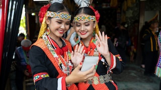 Two people in striking red and black clothing with colorful embellishments stand side by side, looking at a smartphone, while one person waves at the device's camera.