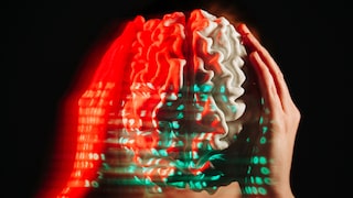 The image shows a white model of a human brain against a black background. A hand holds the brain from the side. Red and turquoise light patterns with numbers and symbols are projected onto the model and the hand. The left side of the image is brightly illuminated in red, while larger white areas of the brain model remain visible on the right side.