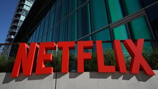 Red Netflix logo in front of the glass facade of a modern building under a blue sky.