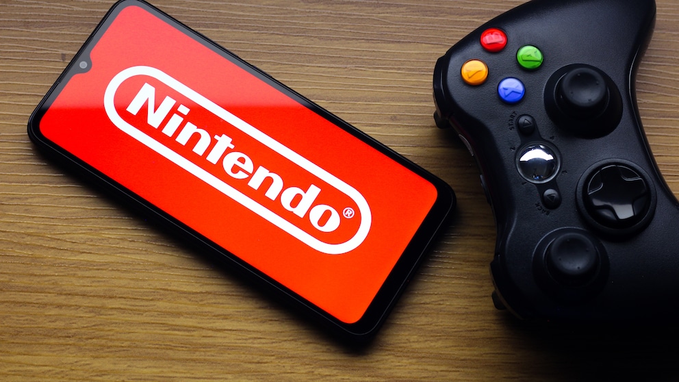 A smartphone with a red Nintendo logo sits on a wooden table. Next to it is a black game controller with colored buttons.