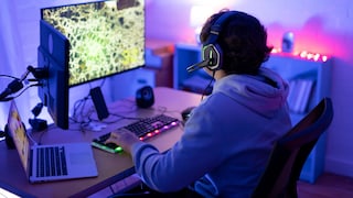 In the image, a person wearing a headset is seated at a desk in front of a large monitor, seemingly playing on a PC. The scene is primarily bathed in blue and violet light. In front of the person are an illuminated keyboard and additional computer accessories. To the left, there is an open laptop. Furniture and additional light sources can be seen in the background.