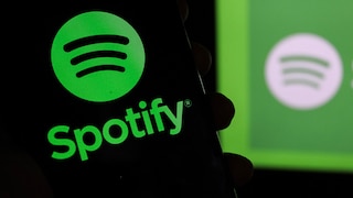 In the foreground of the image, a smartphone displays the green Spotify logo and the word "Spotify." In the background, another blurred Spotify logo is visible against a green surface. The overall color scheme is predominantly black and vibrant green.