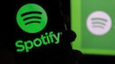 In the foreground of the image, a smartphone displays the green Spotify logo and the word "Spotify." In the background, another blurred Spotify logo is visible against a green surface. The overall color scheme is predominantly black and vibrant green.