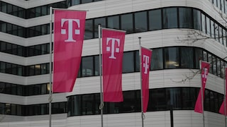 The image shows several magenta Telekom flags with the white "T" logo in front of a modern office building. More flags are visible on the right edge of the image, with a bright facade featuring large dark windows in the background.
