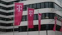 The image shows several magenta Telekom flags with the white "T" logo in front of a modern office building. More flags are visible on the right edge of the image, with a bright facade featuring large dark windows in the background.