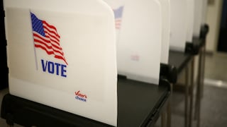 Several white voting booths in a polling station, with the front one displaying a U.S. flag and the words "VOTE" and "Voter's Choice."
